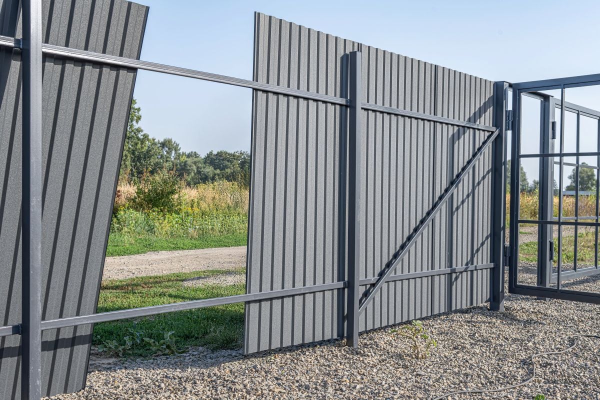 A partially installed corrugated metal fence with an open gate stands on a gravel-covered property